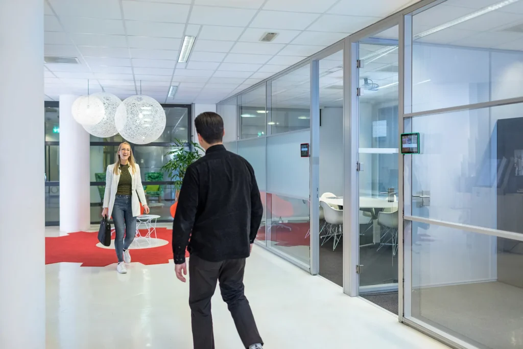 Twee mensen lopen door een moderne kantoorruimte met glazen wanden en grote hanglampen.