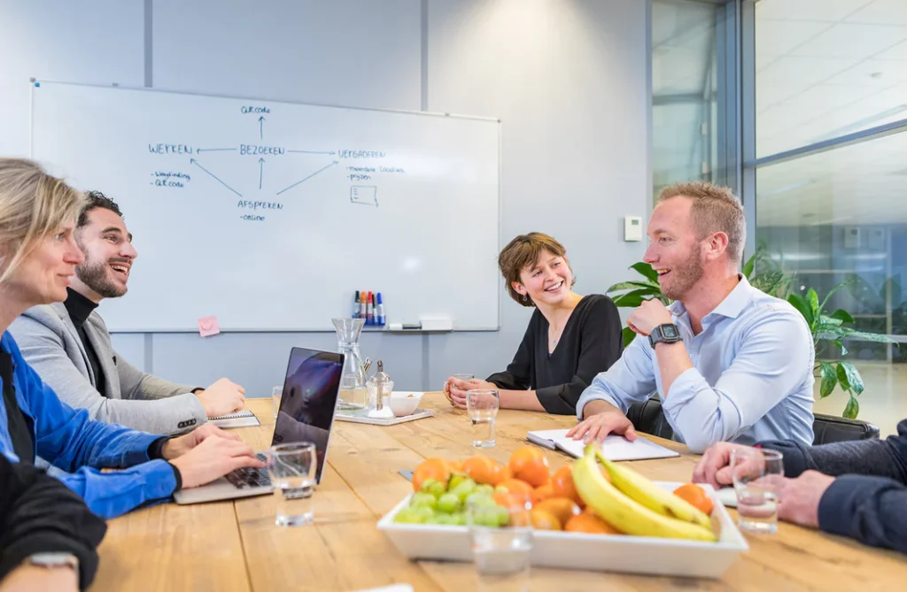 Mensen zitten rond een vergadertafel terwijl ze lachen en praten, met een fruitschaal op de voorgrond en een whiteboard op de achtergrond.