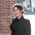 Een vrouw in een zwart colbert poseert buiten tegen een achtergrond van bakstenen en moderne architectuur.